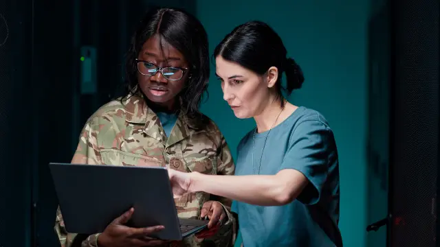 Image of two woman wearing military outfits and looking at a laptop screen together