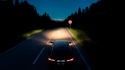Image of a car driving along a rural highway at night, lighting the road ahead as well as a sign up ahead with its front lights