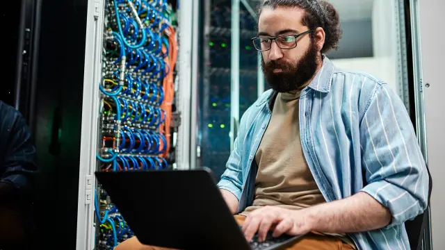 Image of bearded man next to a server cabinet, working on his laptop