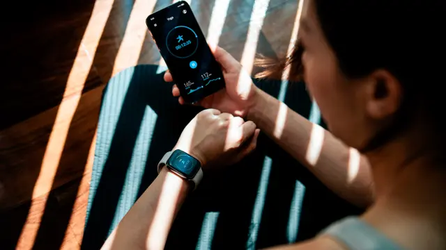 Foto einer jungen Frau, die auf einer Yogamatte sitzt und auf ihre Smartwatch sowie ihr Smartphone schaut