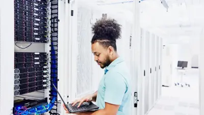 An IT technician works with a laptop in a brightly lit server room. He is standing next to an open server rack with visible network cables and hardware components.