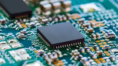 Ein schwarzer Mikrochip mit zahlreichen Anschlussbeinchen ist zentral auf einer grünen Leiterplatte mit vielen kleineren SMD-Bauteilen platziert.